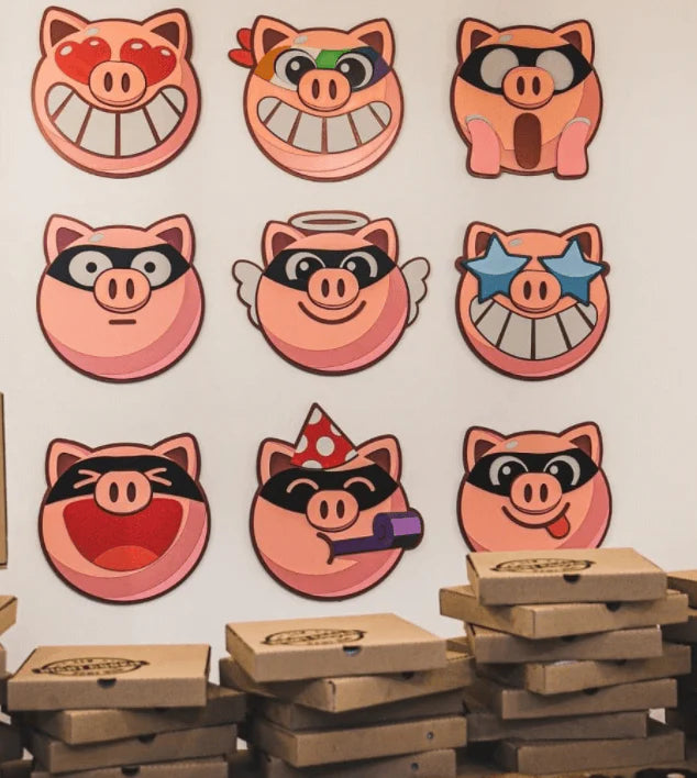 Nine circular pig face stickers or decals with different cartoon expressions are arranged on a white wall above stacked pizza boxes. The pigs wear various accessories like sunglasses, wings, a party hat, and a megaphone.