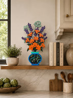 A colorful 3D wooden floral arrangement in a blue patterned vase, styled on a wooden shelf next to books, a small potted plant, and kitchen items in a warm, modern kitchen setting.