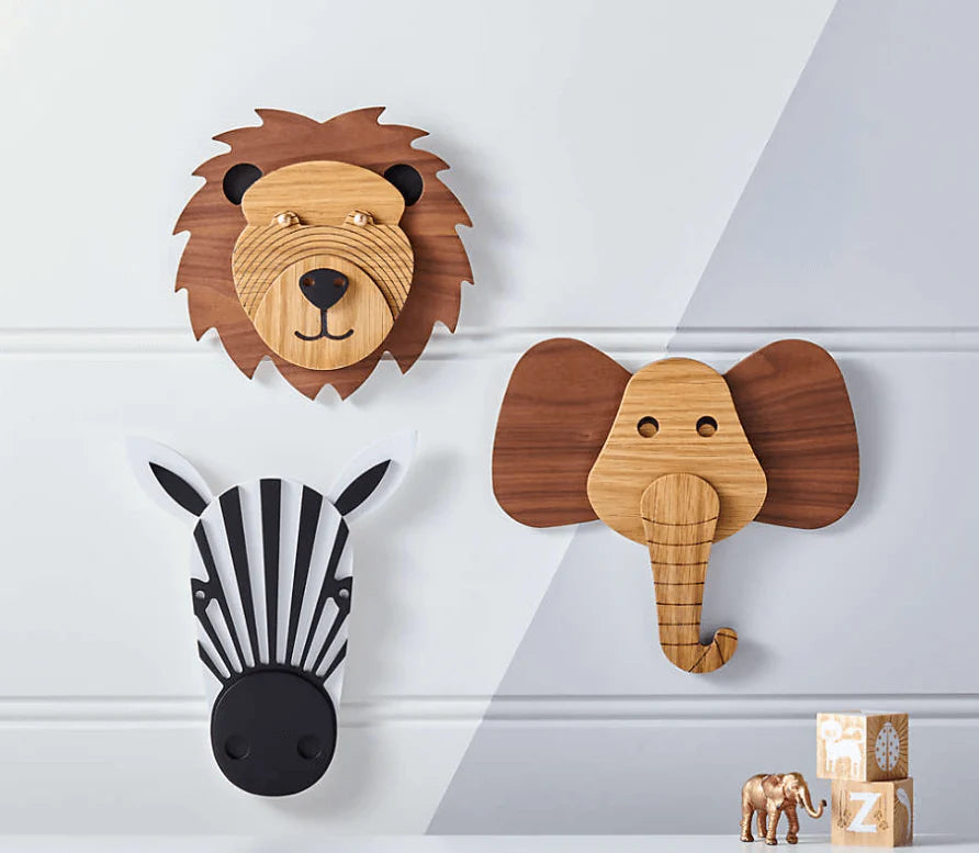Three wooden animal head wall hooks displayed on a light gray paneled wall: a lion, an elephant, and a zebra. Small wooden blocks are placed near the bottom right.
