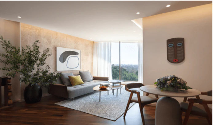 A modern living room interior with a gray sofa, patterned area rug, round coffee table, dining table with chairs, large potted plant, textured accent wall, and a decorative wooden wall piece. No people or animals are present.