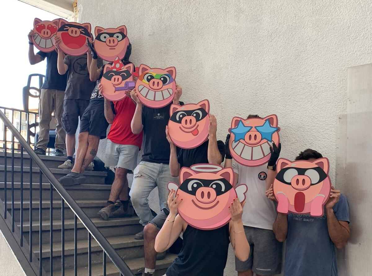 A group of people standing on outdoor concrete stairs holding up large, colorful cartoon pig mask faces to block their views. The masks feature various expressions and accessories like sunglasses, a crown, and wings.