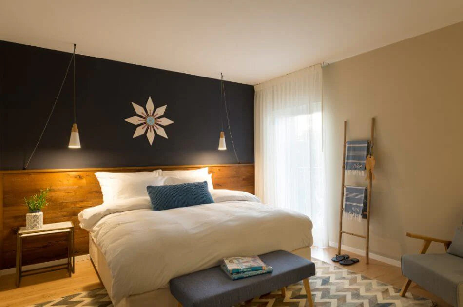 A modern bedroom featuring a bed dressed in white bedding with a blue accent pillow, set against a dark charcoal headboard wall with a white geometric star design. The room has light wood flooring, a patterned rug, a gray armchair, and a blue-and-white striped towel draped over a wooden ladder rack.