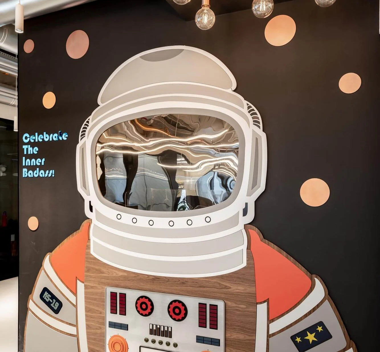 A large astronaut wall cutout decoration in a white spacesuit with orange accents, mounted against a dark wall with decorative polka dots and hanging bulbs.