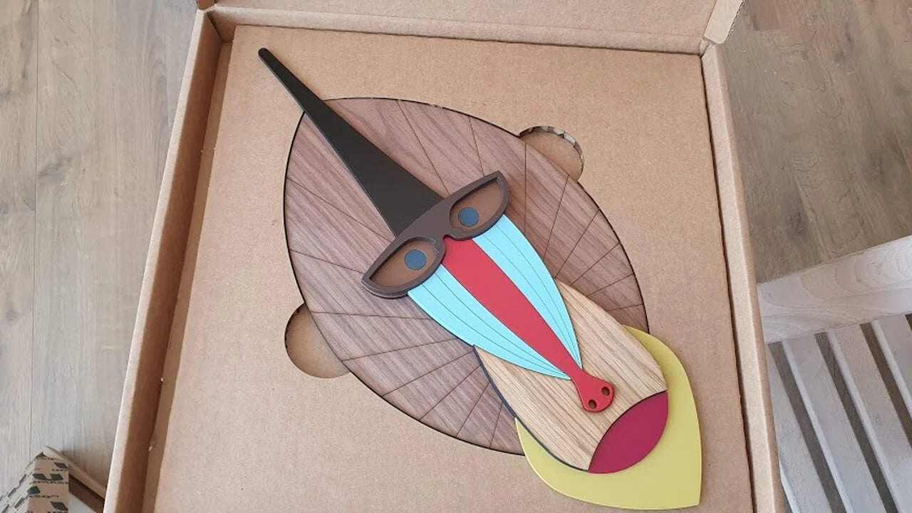 A colorful wooden abstract art piece displayed inside its cardboard packaging box, set on a light wood floor.