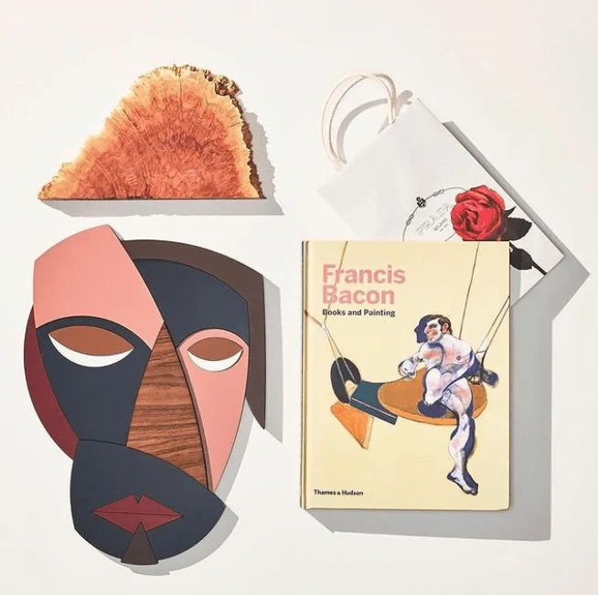 A styled flat-lay featuring a book titled 'Francis Bacon Books and Painting' by Thames & Hudson, alongside decorative art objects including a geometric face mask, a wood slice, and a white gift bag with a red rose, all arranged on a light background.