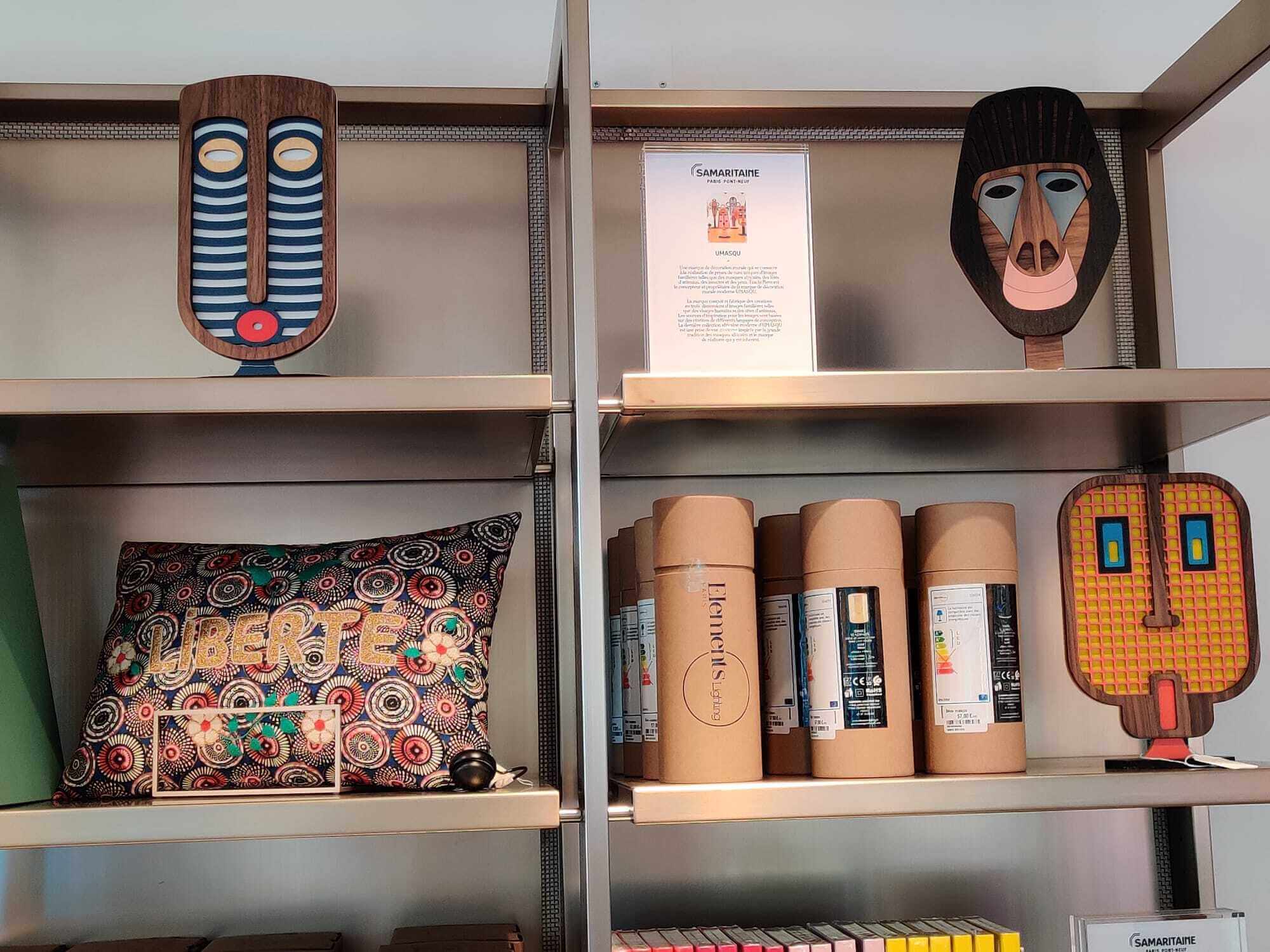 A display of handcrafted Guatemalan artisan products on metal shelving, including two wooden masks, a patterned lumbar pillow, and several cylindrical cardboard boxes of Guatemalan origin.