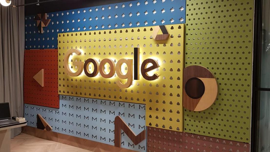 A colorful Google-branded wall display with illuminated Google lettering, geometric panels, and repeating 'M' patterns in a modern interior setting.