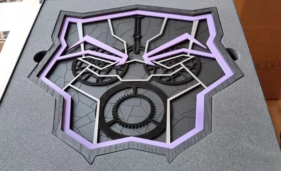 A close-up of a black and purple mechanical emblem or decorative panel mounted on a gray textured backing, with visible inner gear-like details and angular cutout lines.