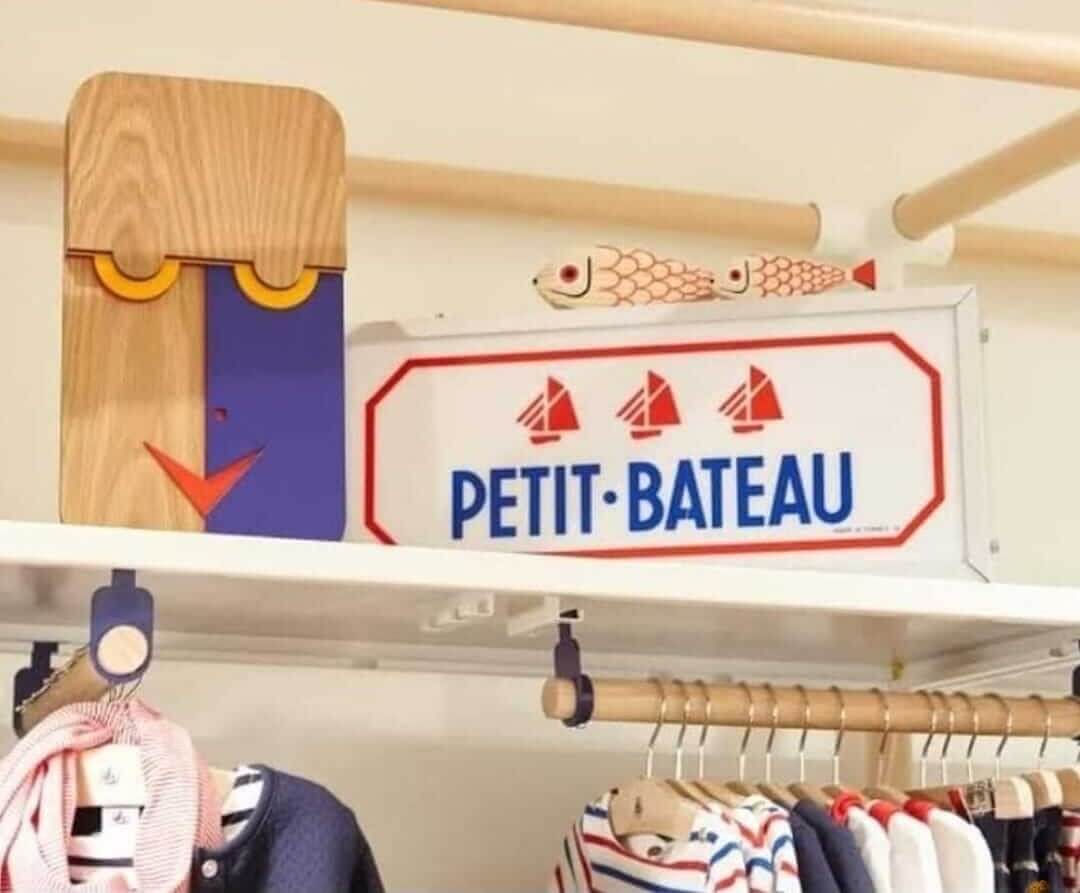 A children's clothing rack with wooden hangers and a white sign reading 'PETIT BATEAU' with red sailboats, shown in a nursery or children's room setting.